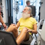 Older man exercising in a gym with a coach guiding him — staying active and managing joint pain after 50